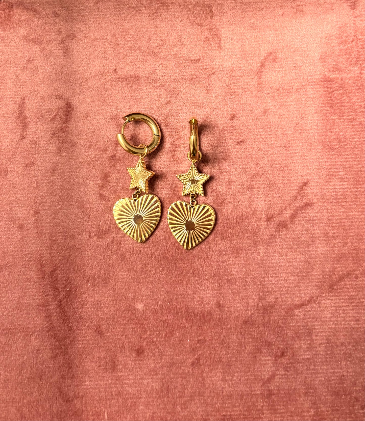 "Twinkle & Trouble" oorbellen – goudkleurige oorringen met een stervormige hanger en een geribbeld hartje met kristal in het midden, gefotografeerd op een zachte roze fluwelen ondergrond
