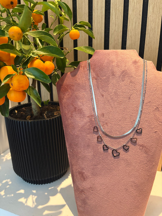 Gelaagde zilveren ketting “Love me in layers” met speelse hartjesbedels, gepresenteerd op een roze display