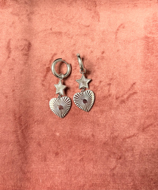 "Twinkle & Trouble" oorbellen – Zilverkleurige oorringen met een stervormige hanger en een geribbeld hartje met kristal in het midden, gefotografeerd op een zachte roze fluwelen ondergrond