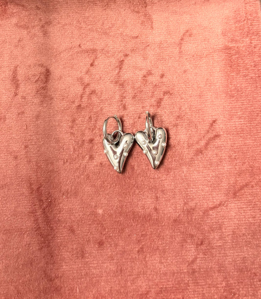 Zilverkleurige hartvormige oorbellen met parelachtige details, gefotografeerd op een roze fluwelen ondergrond