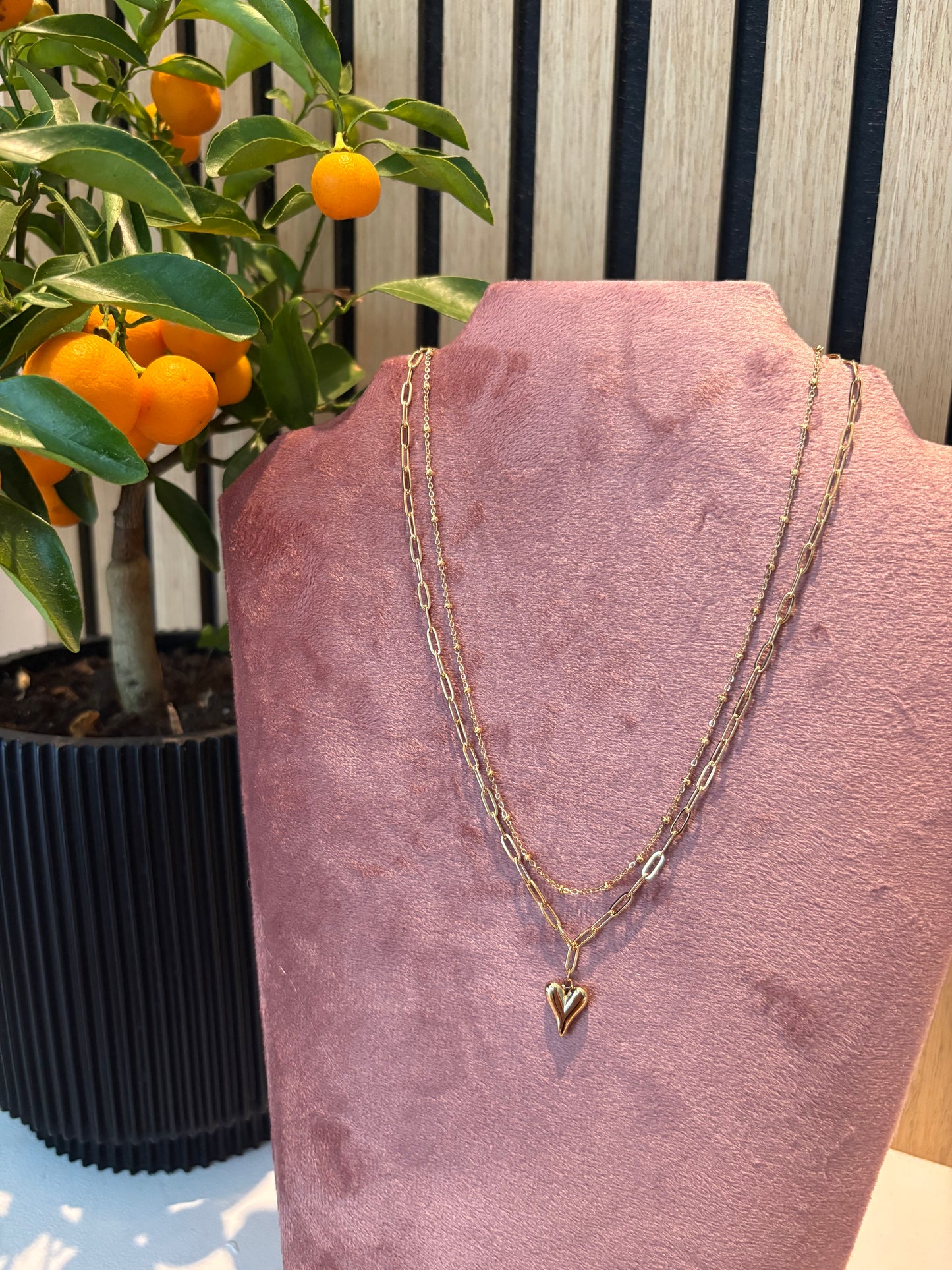 "Chain Reaction" ketting β goudkleurige dubbele ketting met hartvormige hanger, gepresenteerd op een roze fluwelen kettingdisplay