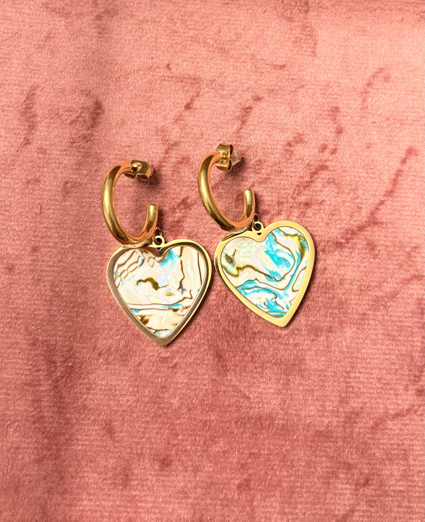 “Mood Swings” oorbellen – goudkleurige hoops met een swirl van goud, witte en blauwe marmeren hartvormige bedel, gepresenteerd op een roze fluwelen achtergrond