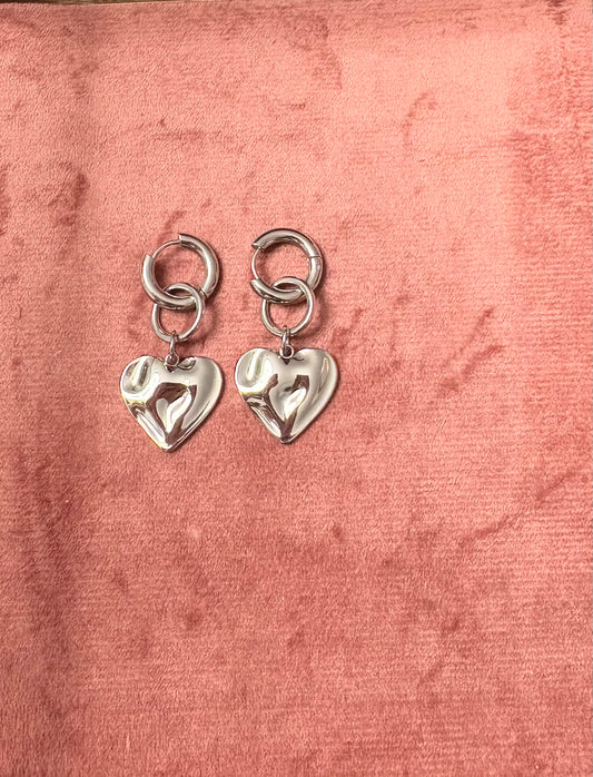 "Wavy Baby" oorbellen – Zilverkleurige hartvormige hangertjes met een golvend reliëf, bevestigd aan ronde oorringen, gepresenteerd op een roze fluwelen achtergrond