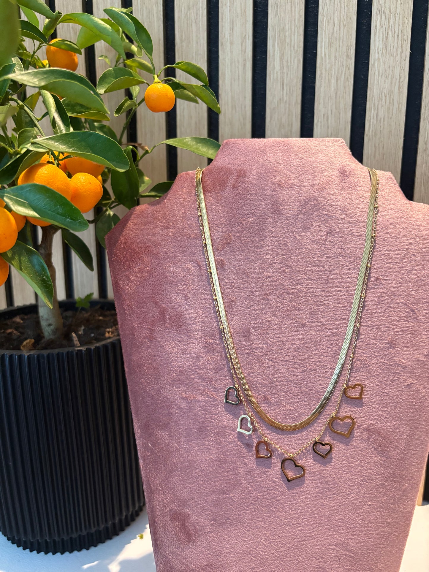 Gelaagde gouden ketting “Love me in layers” met speelse hartjesbedels, gepresenteerd op een roze display