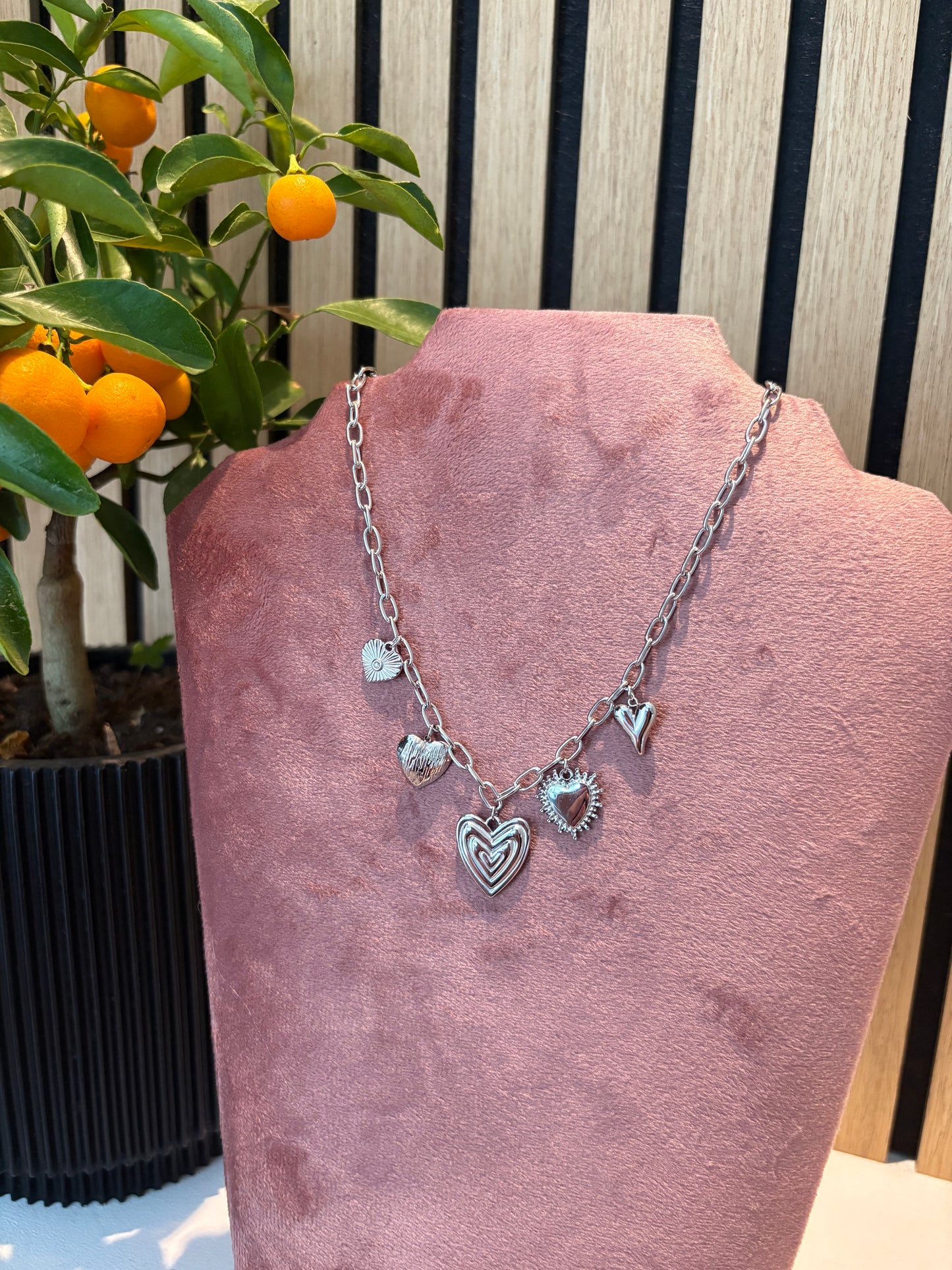"Charmes & Chaos" ketting – zilverkleurige schakelketting met meerdere hartvormige bedels en texturen, gepresenteerd op een roze fluwelen display 