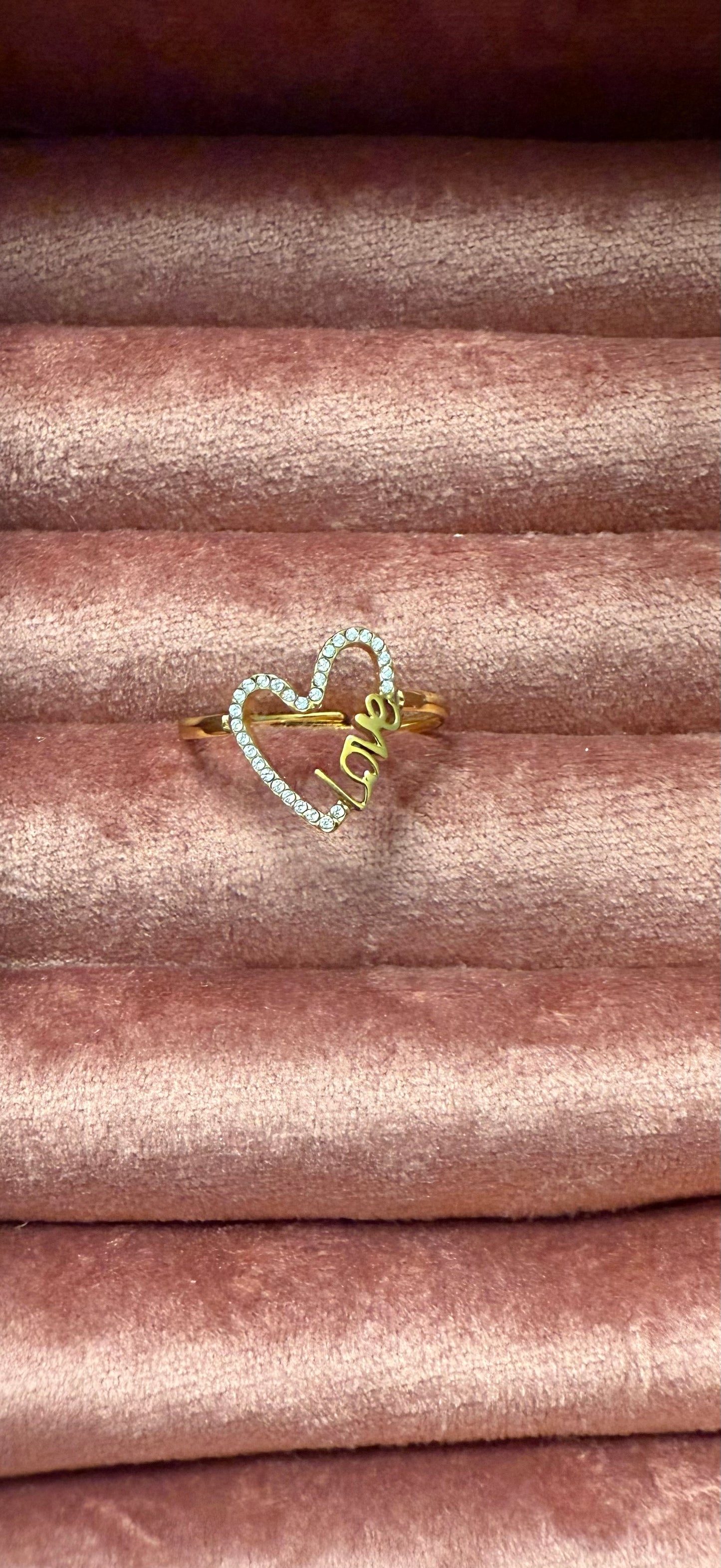 Goudkleurige stainless steel ring met open hartvorm en ingelegde steentjes, met het woord 'Love', gepresenteerd op roze fluwelen sieradendisplay