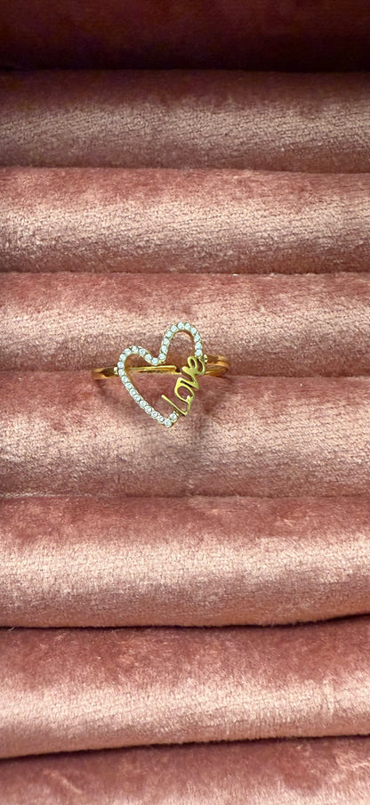 Goudkleurige stainless steel ring met open hartvorm en ingelegde steentjes, met het woord 'Love', gepresenteerd op roze fluwelen sieradendisplay