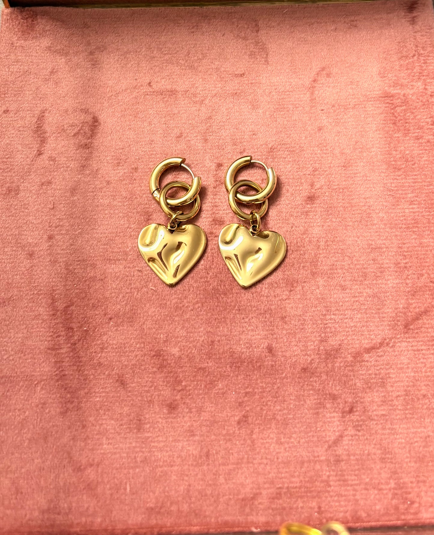 "Wavy Baby" oorbellen – goudkleurige hartvormige hangertjes met een golvend reliëf, bevestigd aan ronde oorringen, gepresenteerd op een roze fluwelen achtergrond