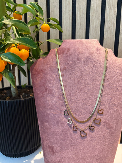 Gelaagde gouden ketting “Love me in layers” met speelse hartjesbedels, gepresenteerd op een roze display 